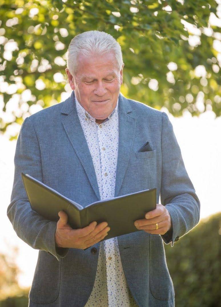 Iain Luck, a distinguished Civil Marriage Celebrant, stands with a warm smile as he reads from a book, against a backdrop of sunlit greenery,